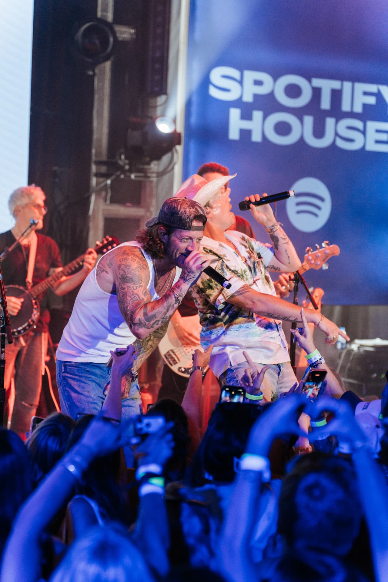 Florida Georgia Line singing side by side at the front of a blue-lit stage in front of a Spotify House backdrop, one in a white tank top and backward camo cap, the other in a cowboy hat and tropical shirt, with a banjo and bass player behind them and a packed crowd raising phones