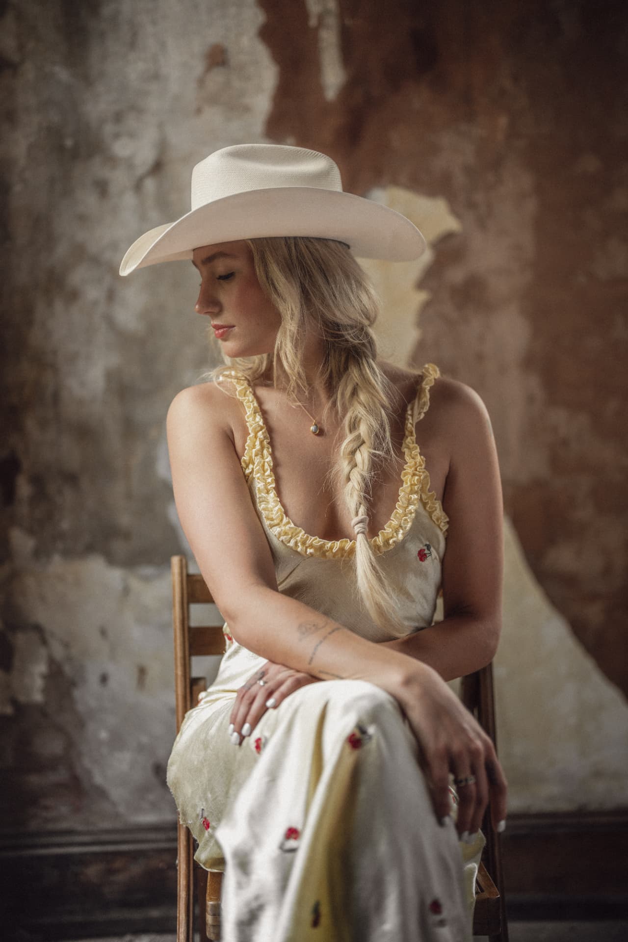 Diner seated on a wooden chair against a peeling wall, wearing a white cowboy hat and a silk slip dress