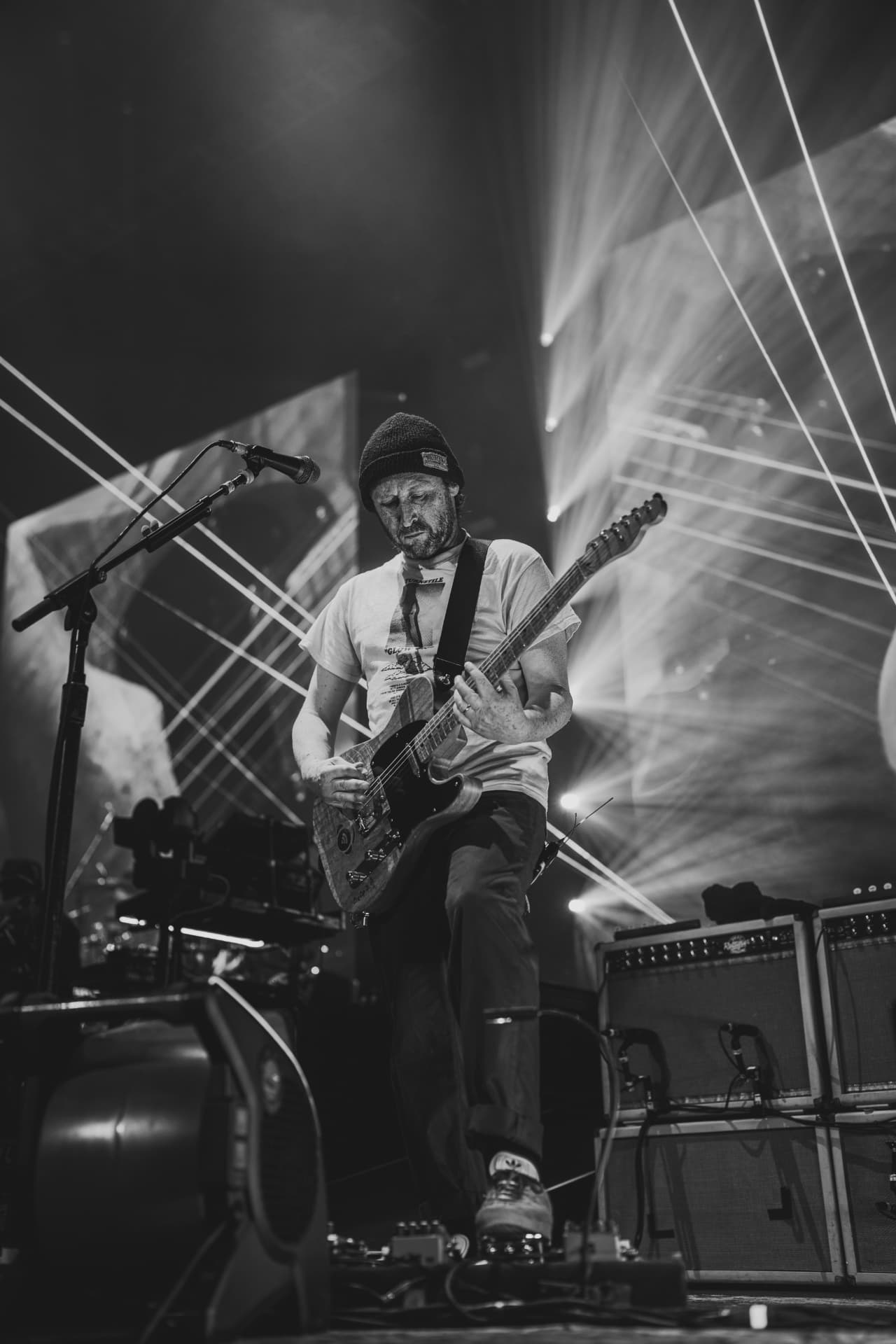 Black-and-white portrait of the guitarist in a beanie playing a Telecaster beside stacked amps under light beams