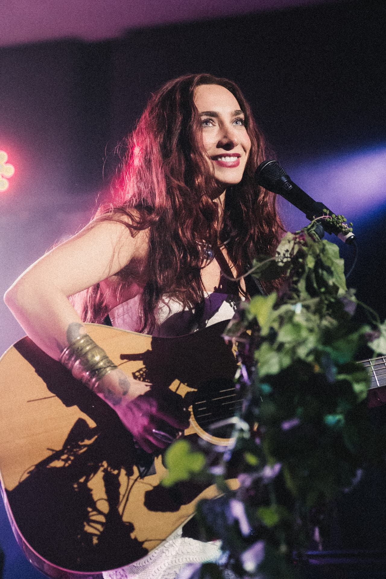 Anna Graves smiling at the microphone with her acoustic guitar under purple and pink stage light, greenery dressed around the stand