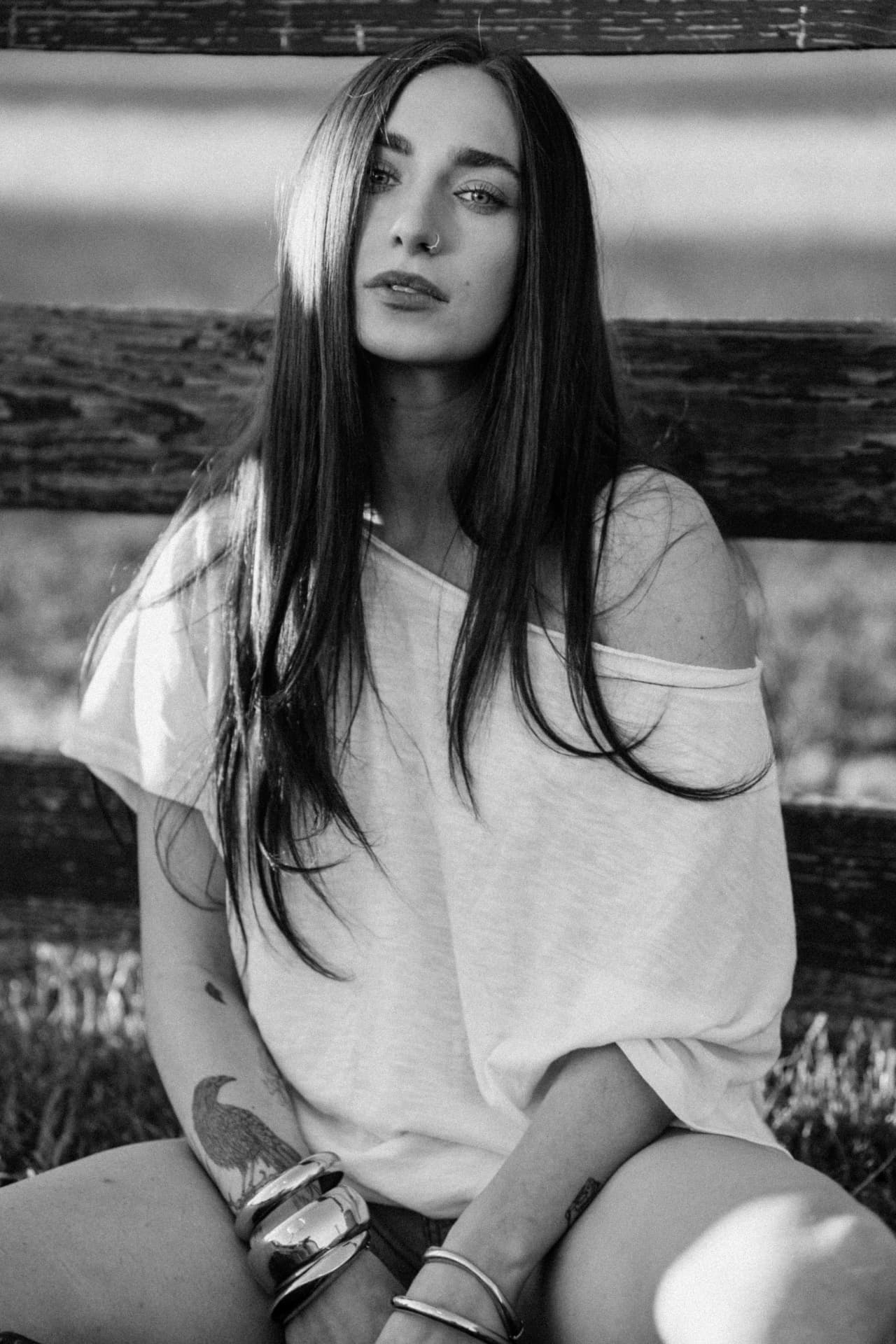 Black-and-white portrait of Anna Graves sitting on the grass in front of a split-rail fence, an off-the-shoulder tee and stacked cuffs, a bird tattoo on her forearm