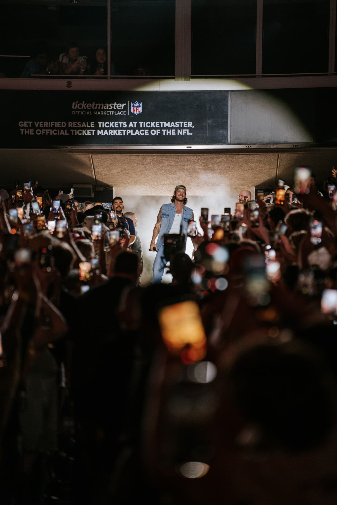 A performer in a sleeveless denim vest and backward camo cap walking onto the field through a stadium tunnel lined with fans holding up phones, glass suites overlooking the corridor above