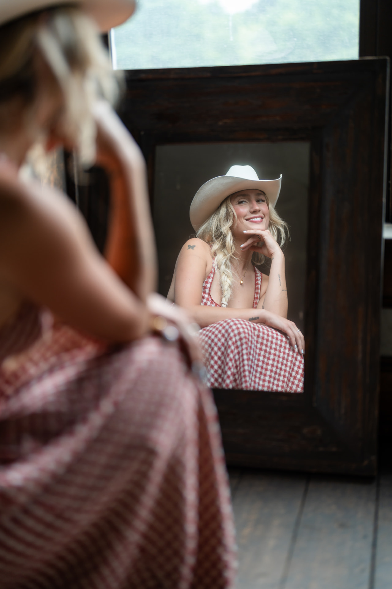 Diner smiling at her reflection in a large dark-framed mirror, wearing a white cowboy hat and a red-and-white checkered dress
