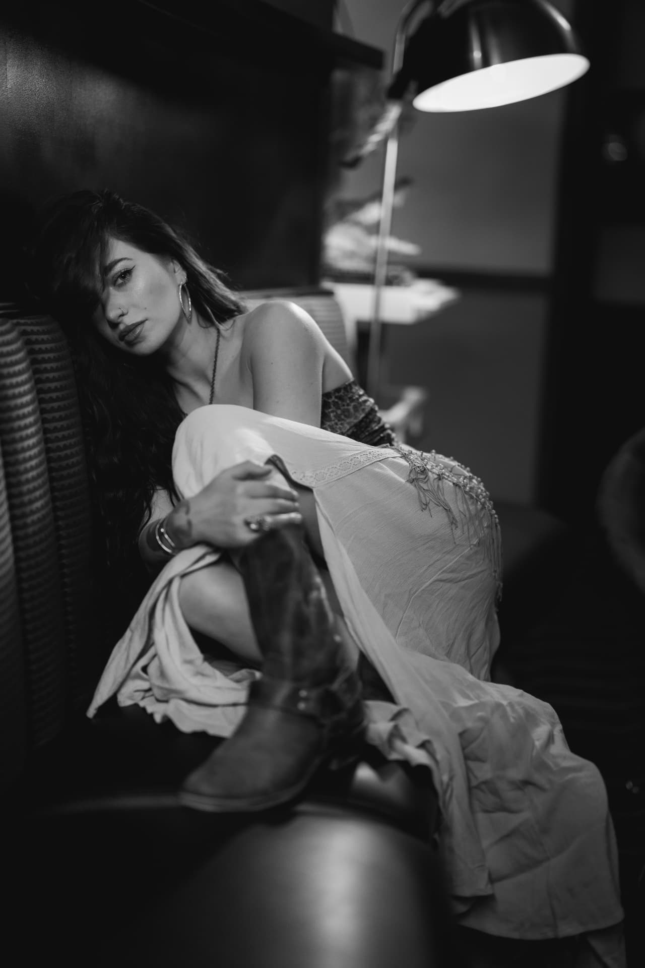 Black-and-white portrait of Anna Graves curled up on a ribbed lounge sofa, a domed lamp glowing over her shoulder