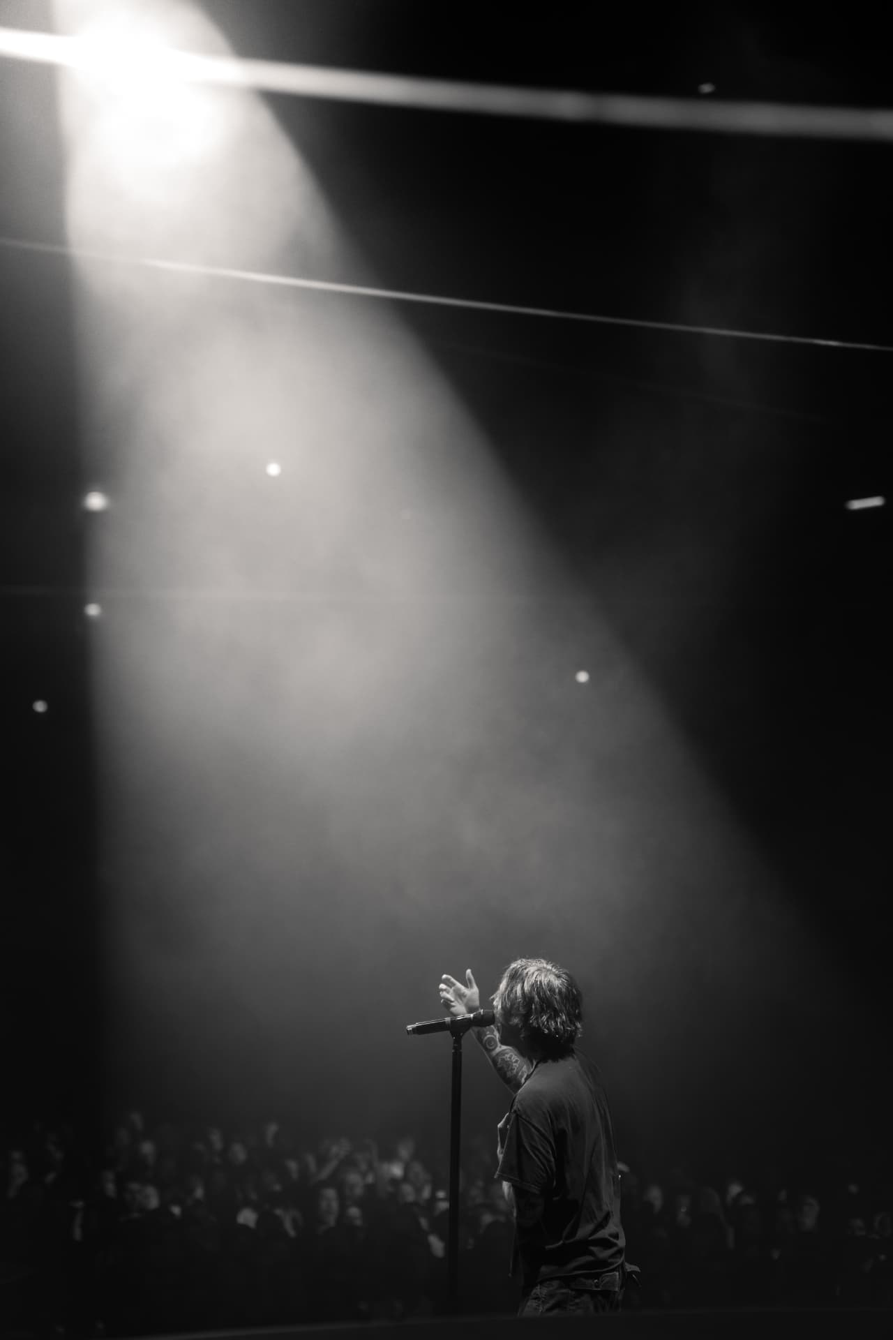 Black-and-white shot of the singer from behind at the mic stand in a wide spotlight beam over a dark crowd