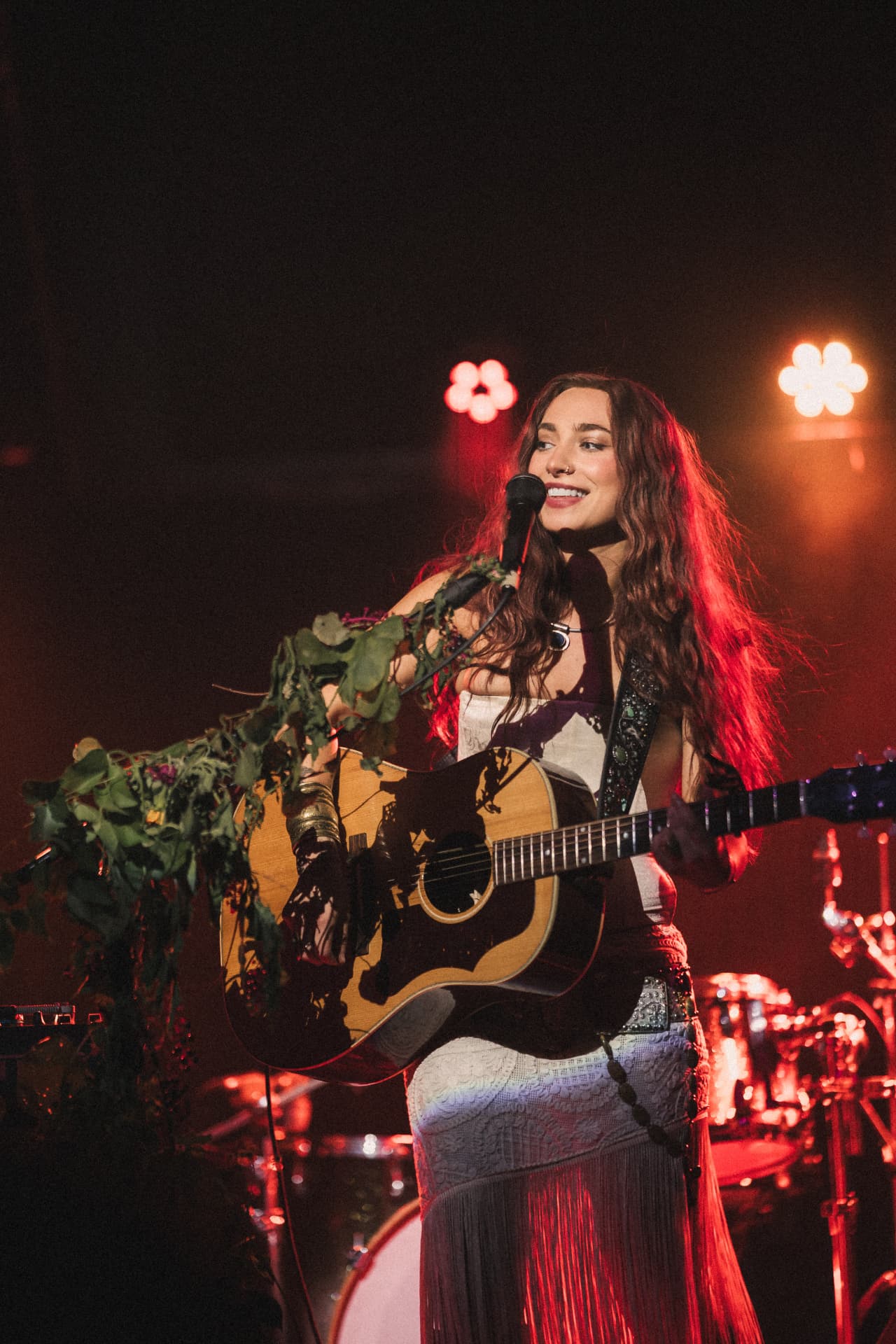 Anna Graves singing with her guitar under warm red stage light, the drum kit glowing behind her