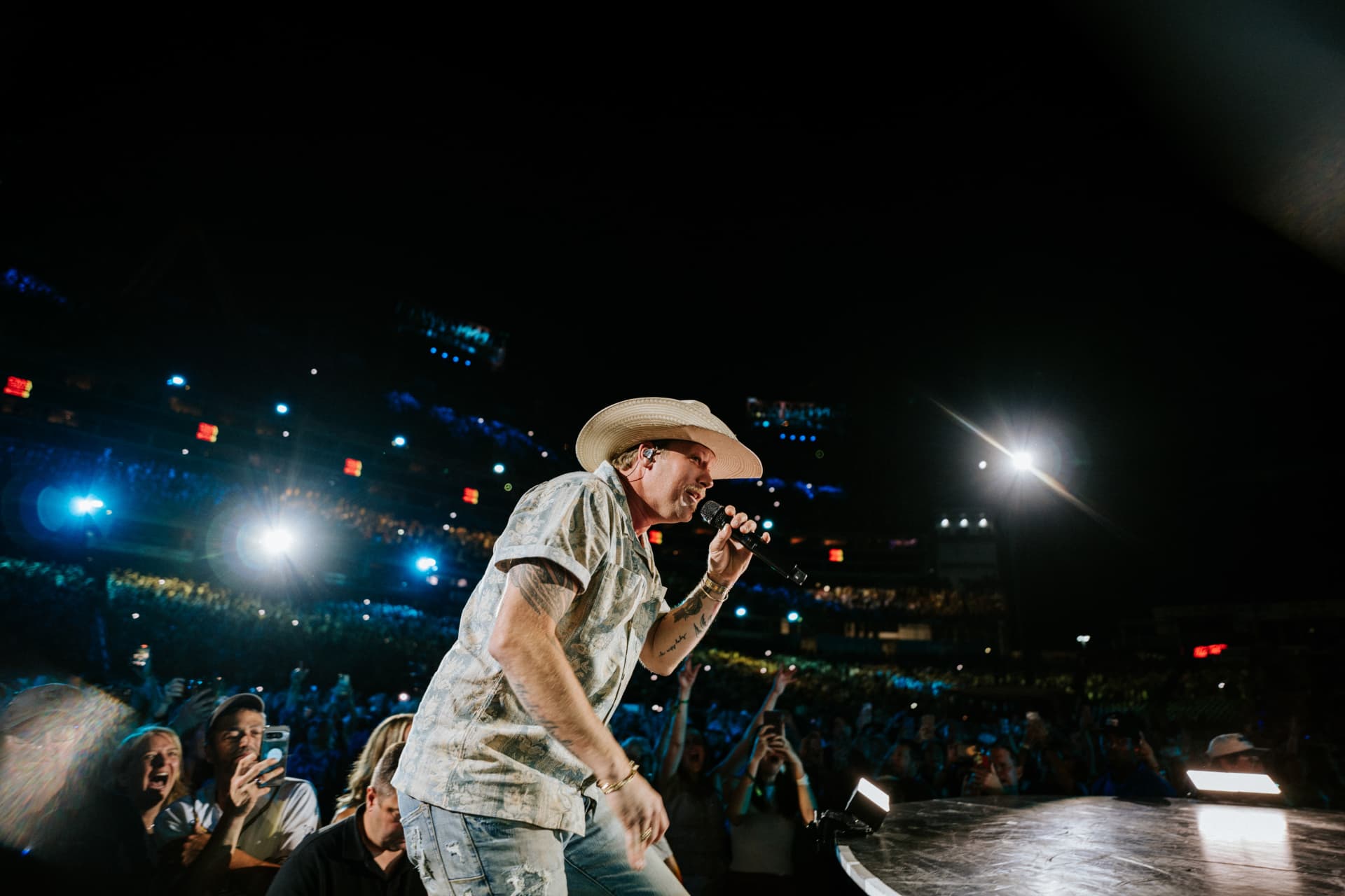 A performer in a straw cowboy hat and patterned shirt singing at the edge of a stage thrust, with a stadium's tiered seating bowls full of fans lit behind him