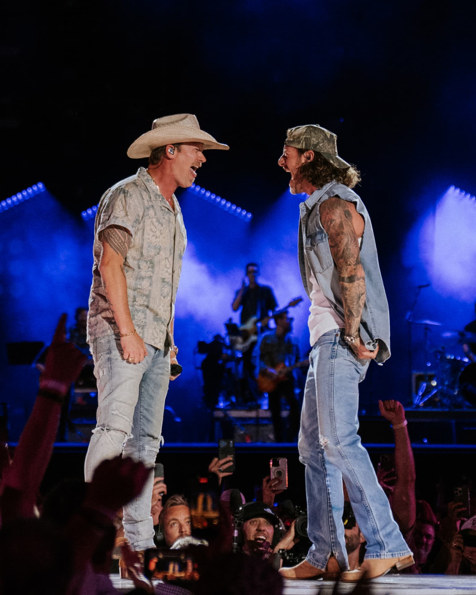 The two performers face to face mid-song on stage, one in a cowboy hat and patterned shirt, the other in a backward camo cap and sleeveless denim vest, blue stage wash and lighting trusses behind them