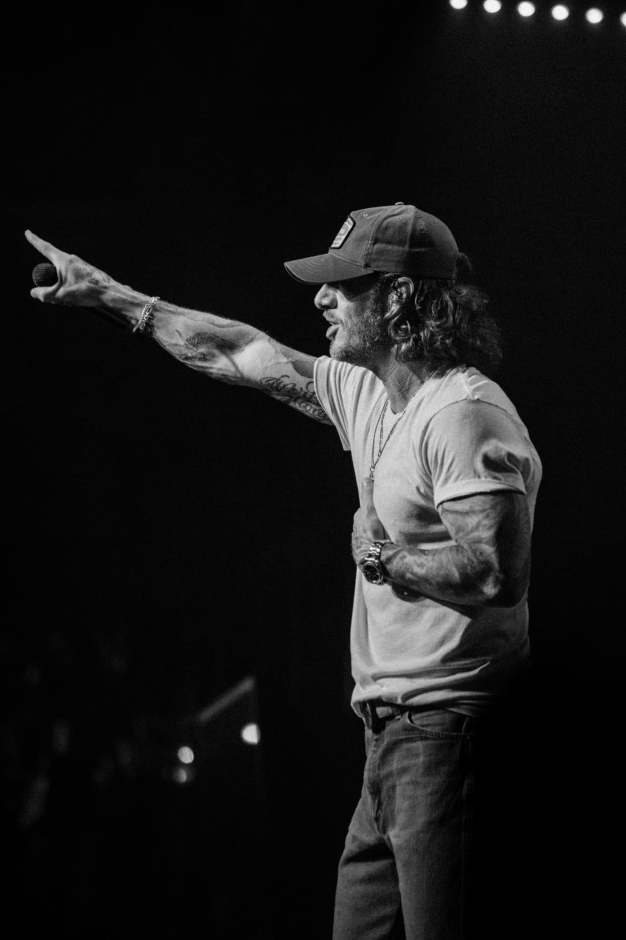 Black-and-white side profile of a performer in a baseball cap pointing out to the crowd, microphone in one hand and the other over his chest, stage lights blurred behind him