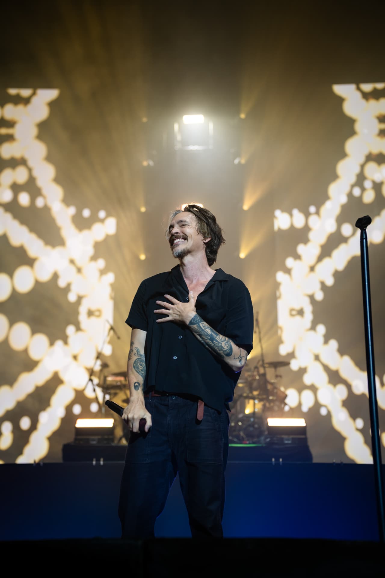 The singer smiling with eyes closed and a hand over his heart mid-song, golden backlight and haze filling the arena stage behind him