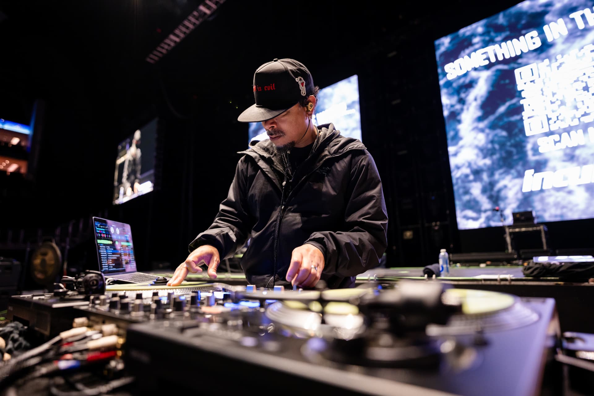 The DJ in a black cap working turntables and a laptop on a dark stage, giant stage screens glowing behind him