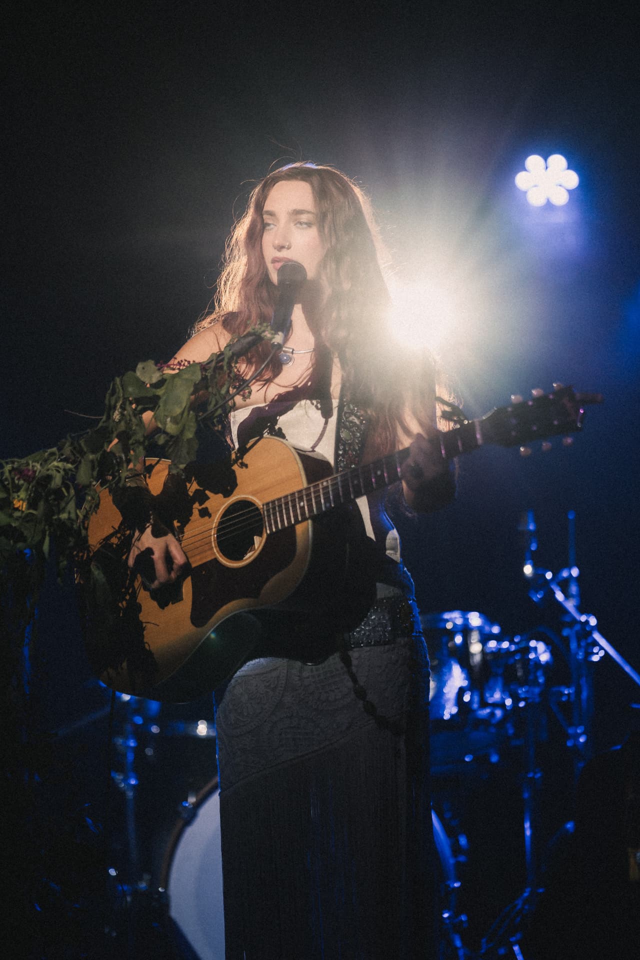 Anna Graves playing acoustic guitar under a bright white backlight on a blue-lit stage