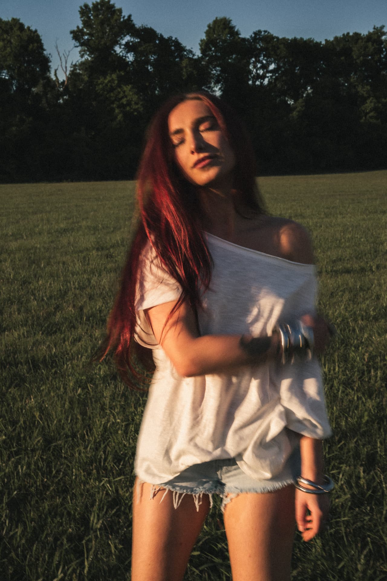 Anna Graves in a white off-the-shoulder tee and cutoff shorts in a mown field at golden hour, her long red hair catching the last light