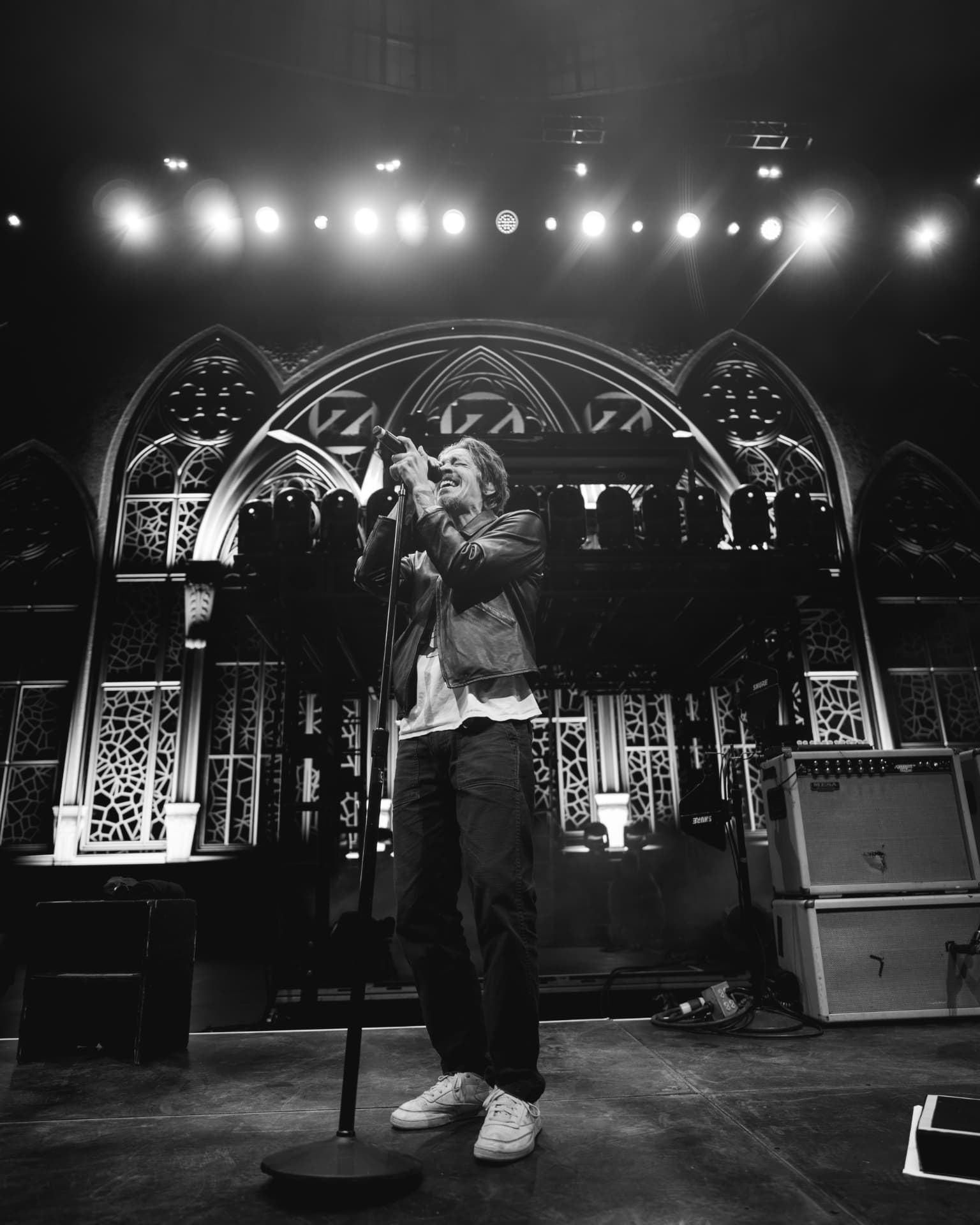 Black-and-white full-length shot of the singer leaning back into a mic on a stand, gothic arch latticework glowing behind him and an amp stack to one side