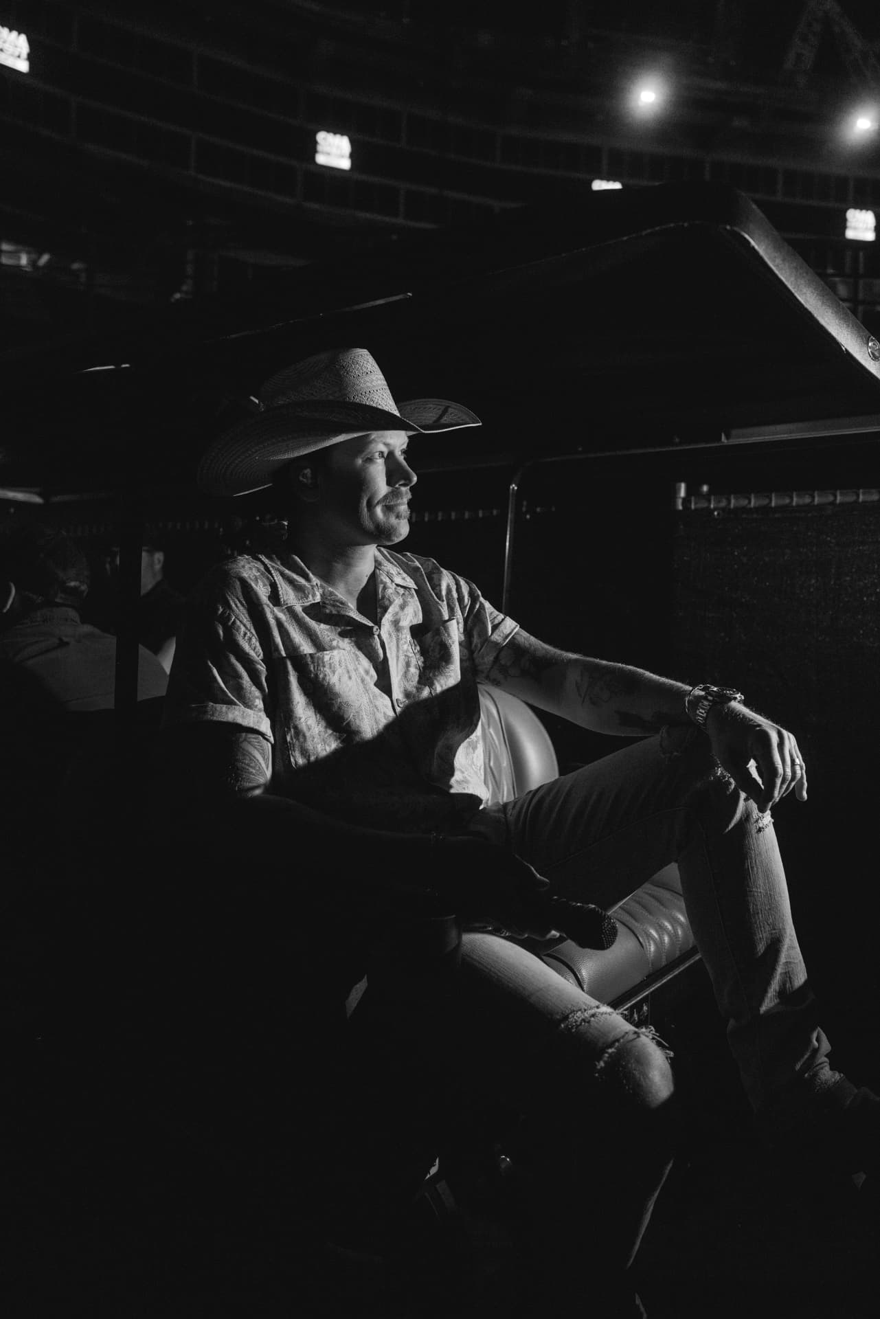 Black-and-white candid of a performer in a cowboy hat sitting in a golf cart with a microphone, stadium concourse and rafters rising behind him