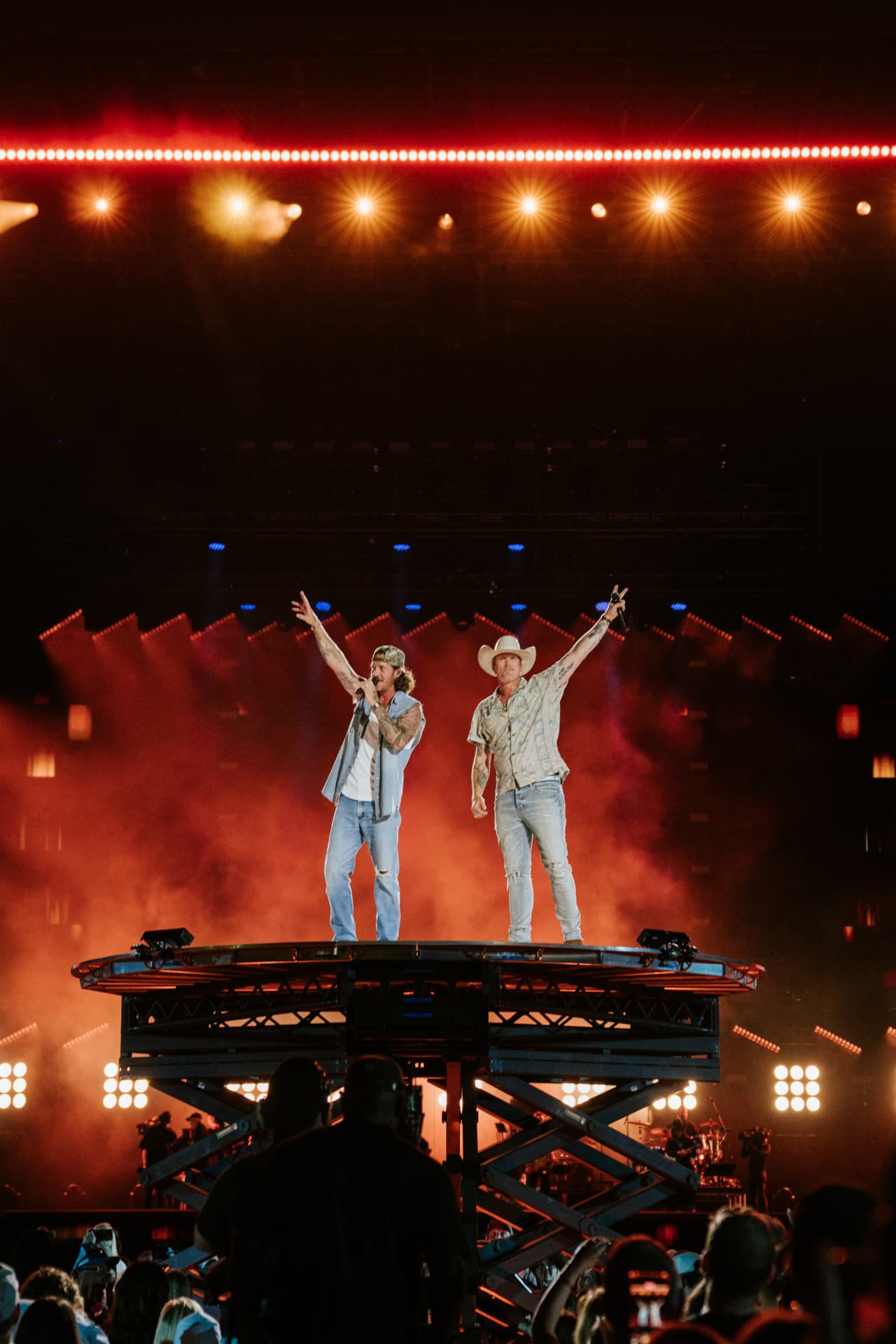 Both performers standing on a raised scissor-lift platform high above the crowd with arms out, lit by red haze and rows of warm spotlights