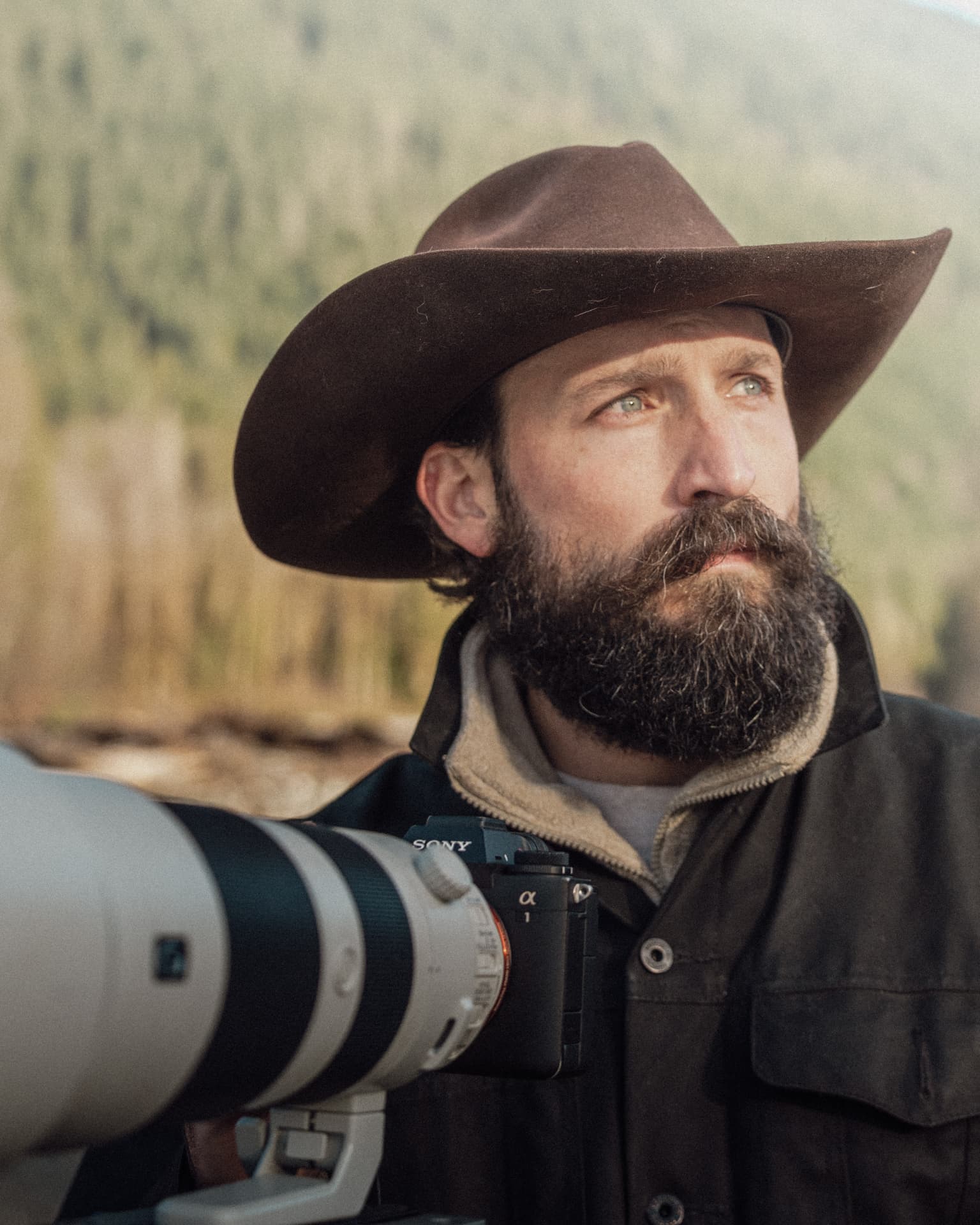 Devin Groody in a cowboy hat looking off-camera behind a camera with a white telephoto lens