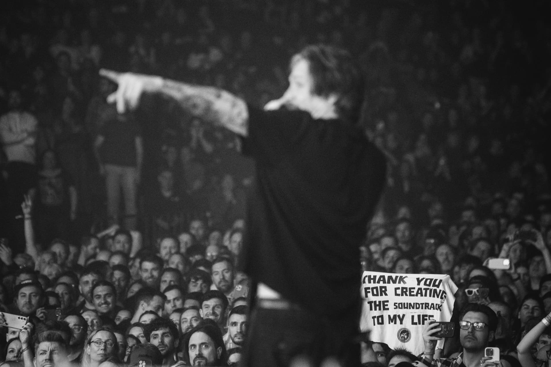 Black-and-white shot from behind the singer pointing out over a packed crowd, a fan holding up a shirt that reads 'Thank you for creating the soundtrack to my life'