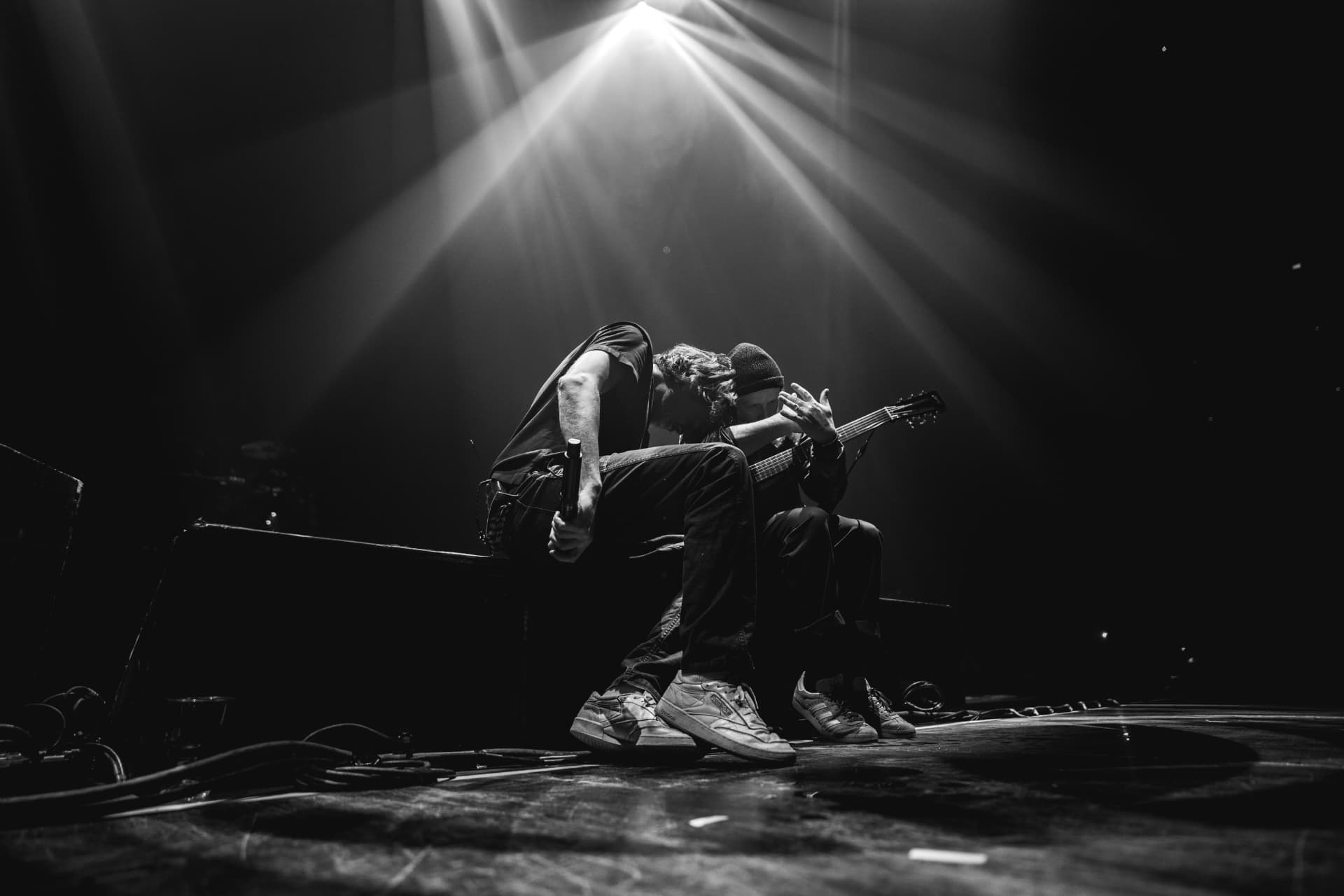 Black-and-white low-angle shot of the singer and guitarist sitting close together on the stage floor under a single spotlight